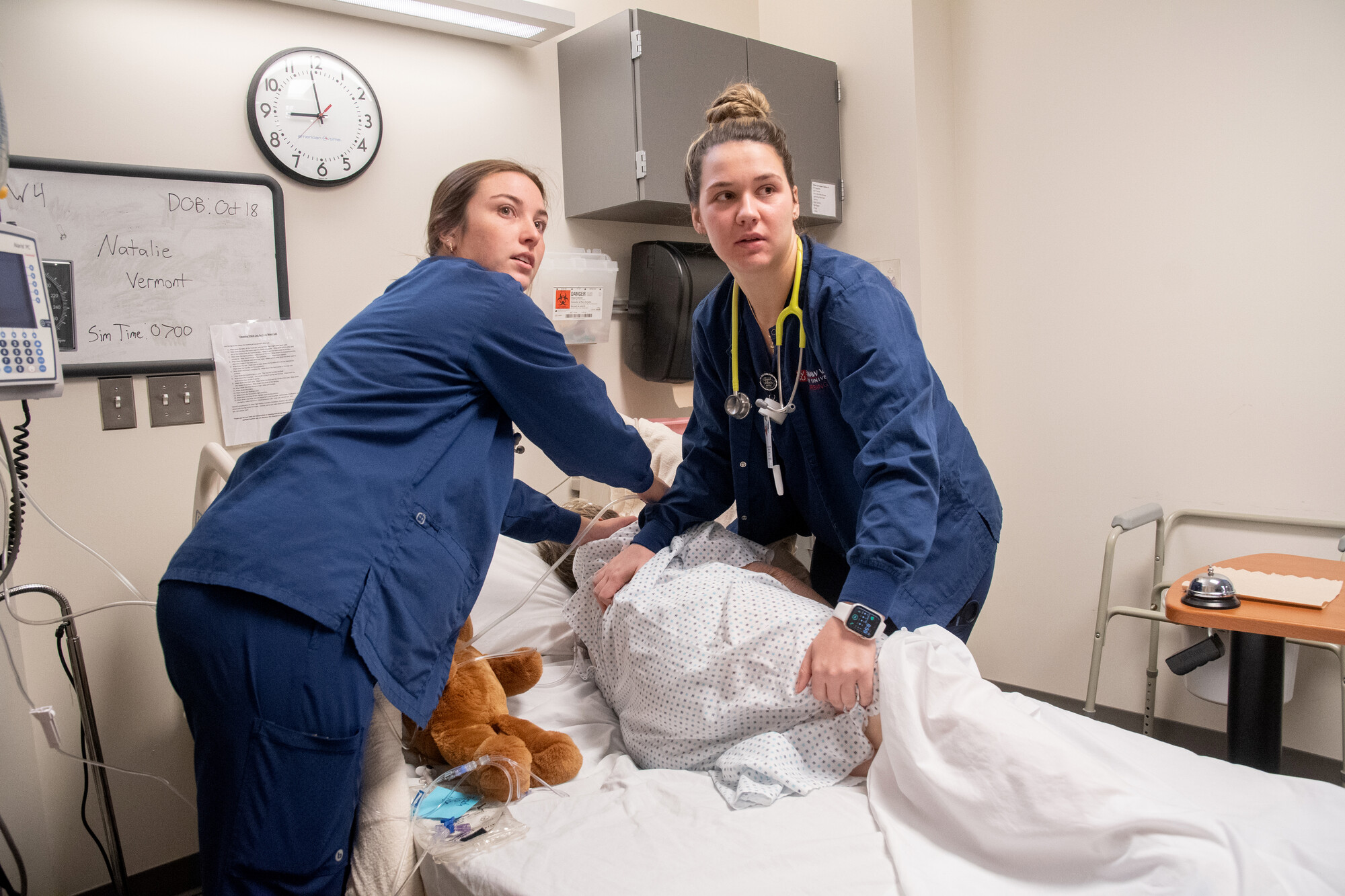 Two young women in blue scrubs work with patient in hospital bed